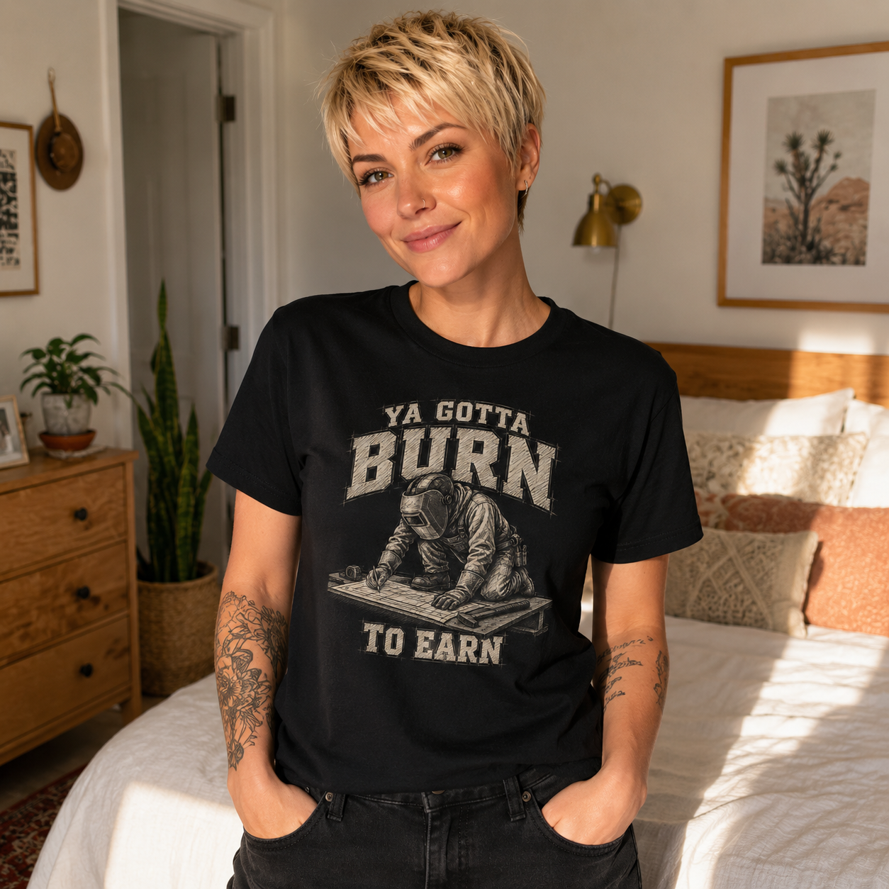 A woman with short blonde hair stands in a bedroom wearing a black graphic T-shirt with the text 'YA GOTTA BURN TO EARN' and a distressed illustration of a person writing at a desk. The room has a bed, dresser, plants, and warm natural light.
