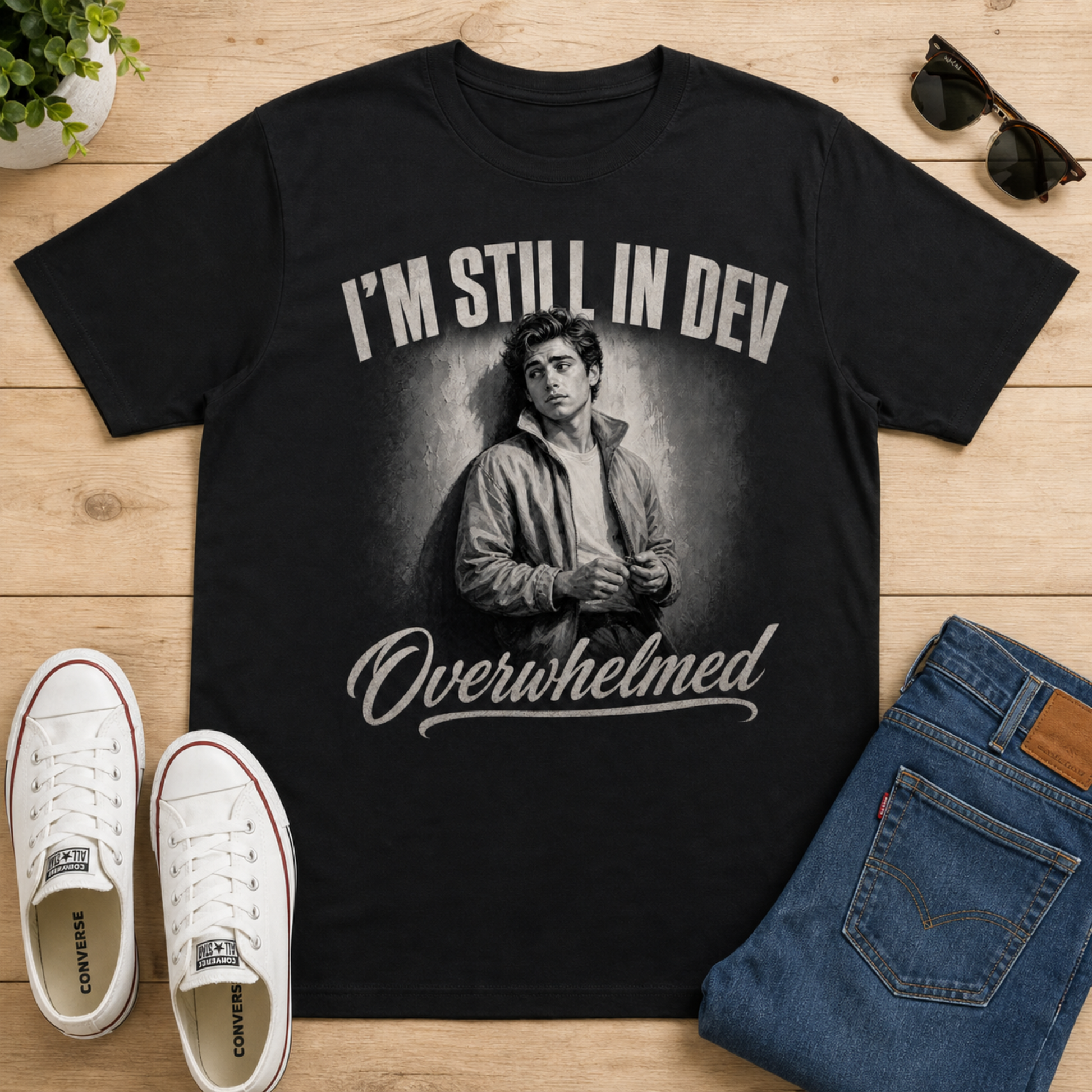 A black graphic t-shirt featuring a grayscale illustration of Jim Jones with the text 'I'M STILL IN DEV' above and 'Overwhelmed' below, styled in a flat lay on a light wood background with white sneakers, blue jeans, and sunglasses.