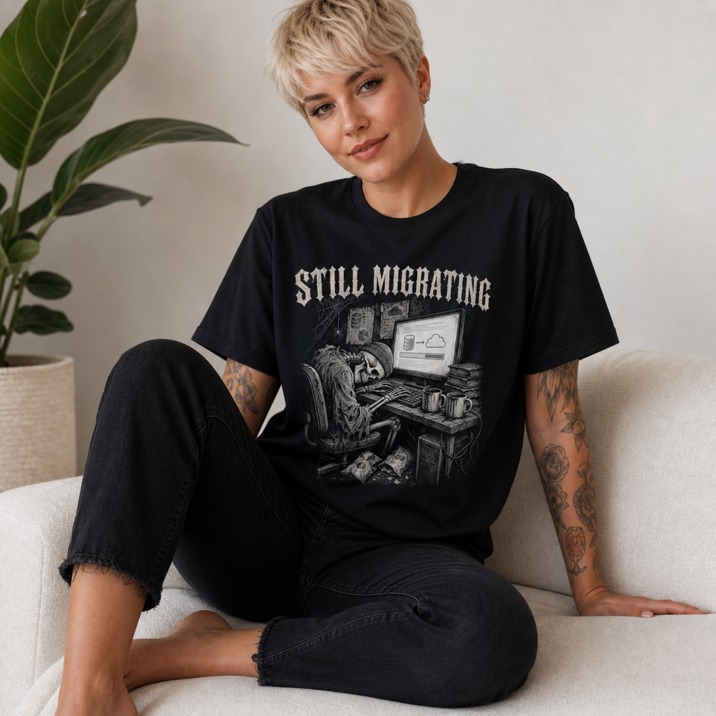 A woman with short blonde hair sits on a light beige couch wearing a black graphic t-shirt with the text 'STILL MIGRATING' and a sketch of a skeleton at a desk. A potted plant is visible in the background.
